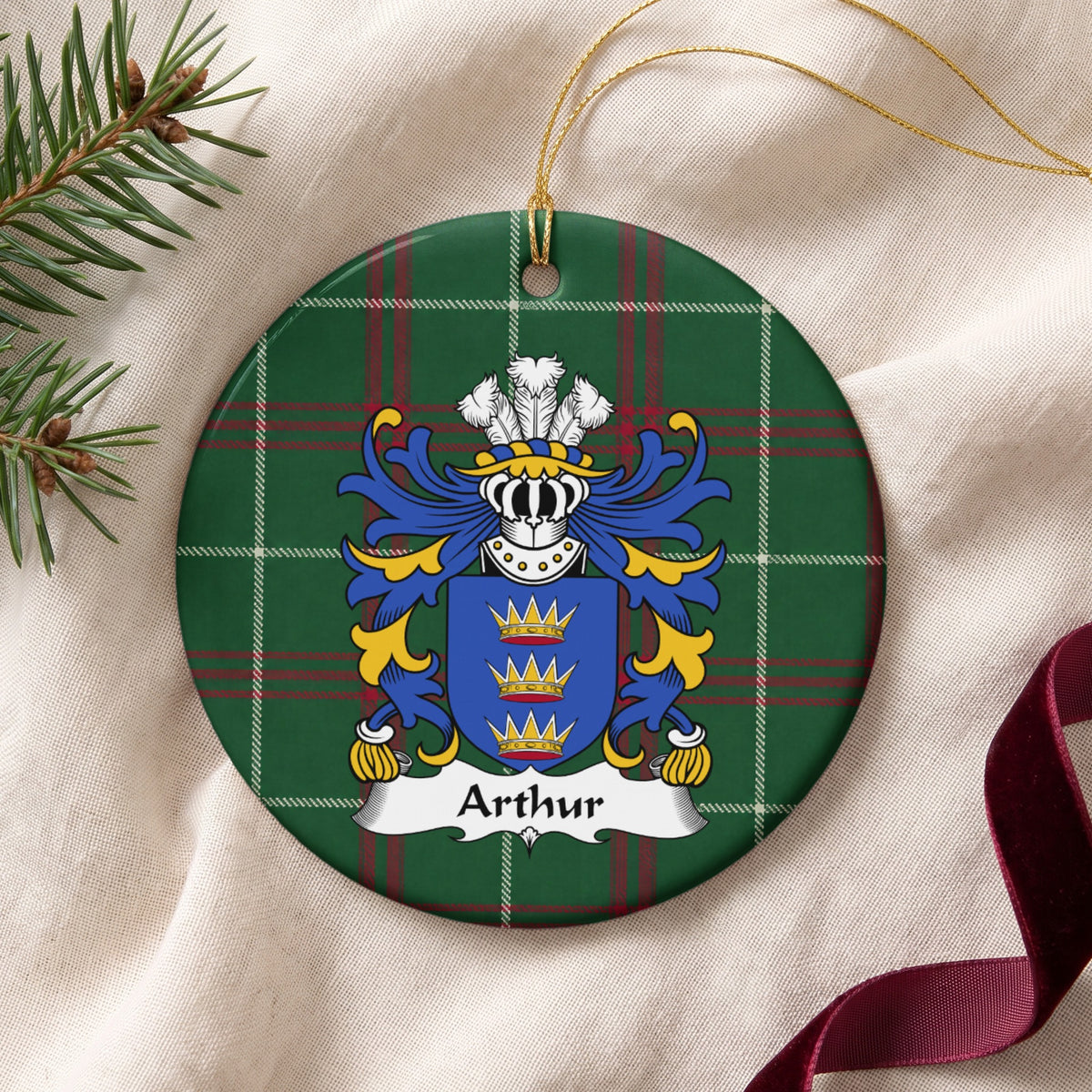 Arthur family crest and surname on a round Welsh national tartan ornament with a gold cord, resting on cream fabric beside an evergreen sprig and burgundy ribbon.