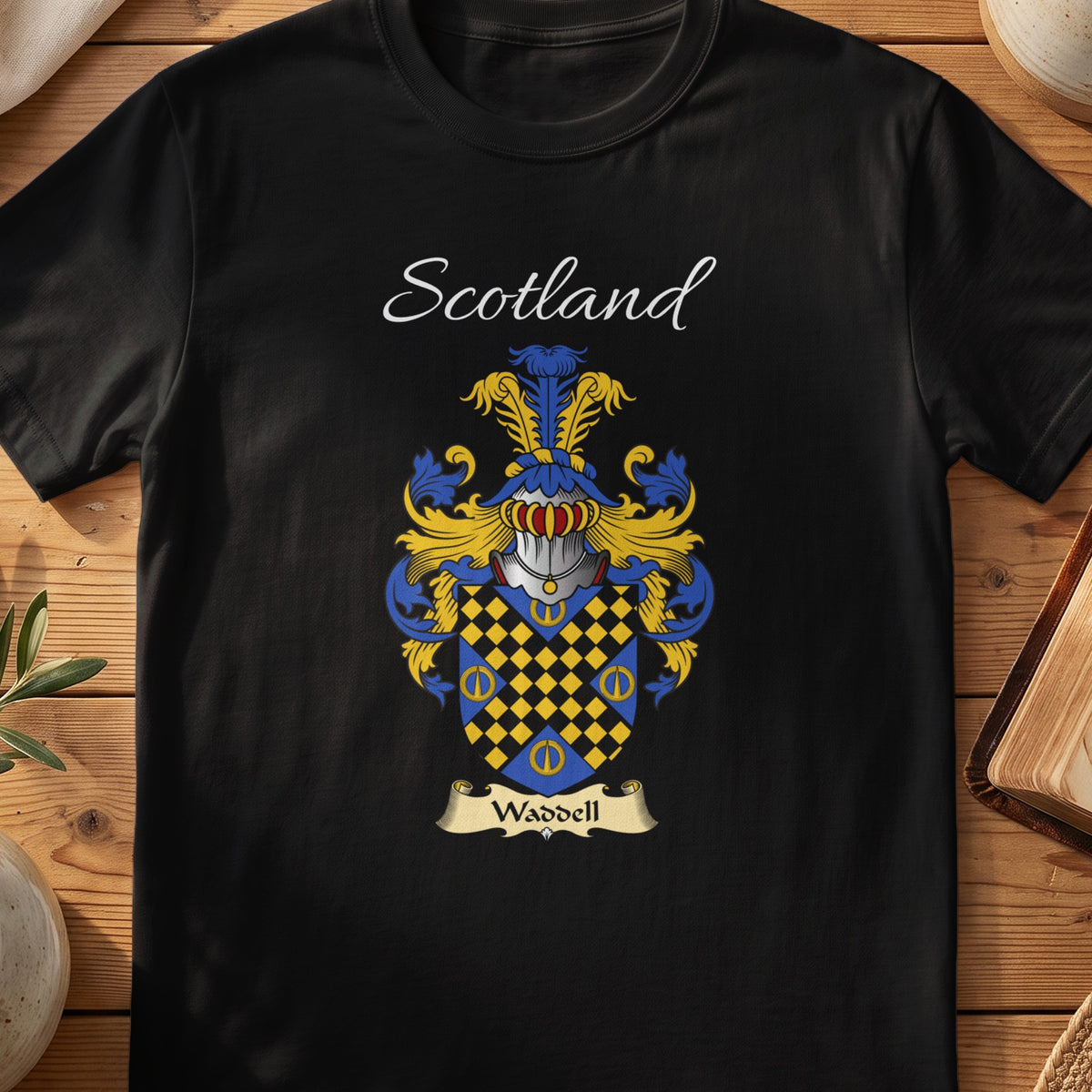 Black Waddell Scottish family crest T-shirt laid flat, showing “Scotland” above a blue-and-gold shield patterned with small chequered diamonds.