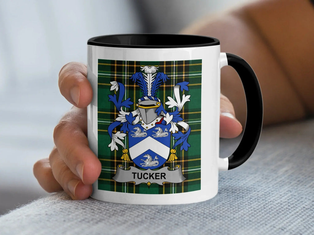 White mug with black rim held by hand, featuring a blue and white coat of arms with "Tucker" banner.