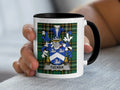 White mug with black rim held by hand, featuring a blue and white coat of arms with "Tucker" banner.