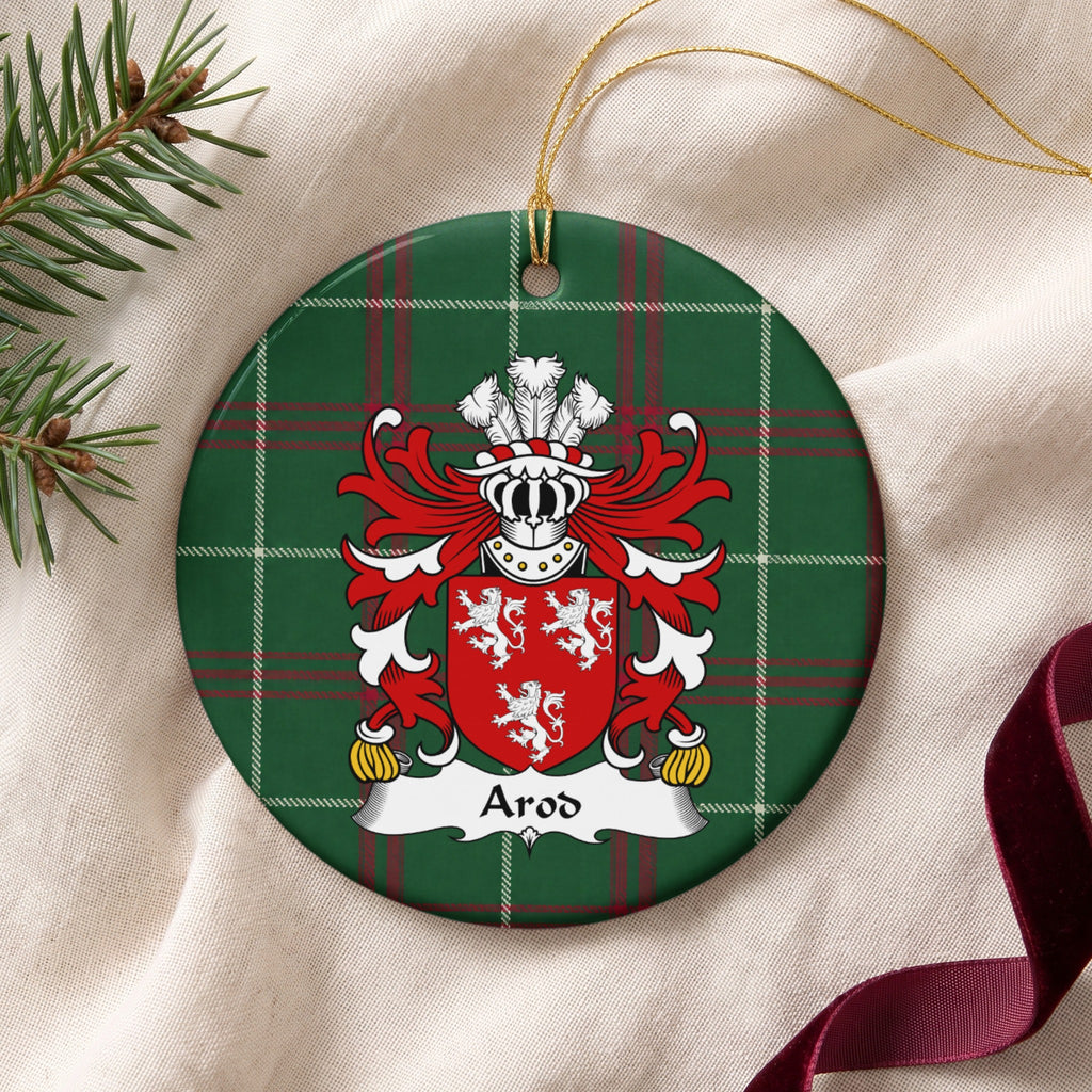 Arod family crest and surname on a round Welsh national tartan ornament with a gold cord, resting on cream fabric beside an evergreen sprig and burgundy ribbon.