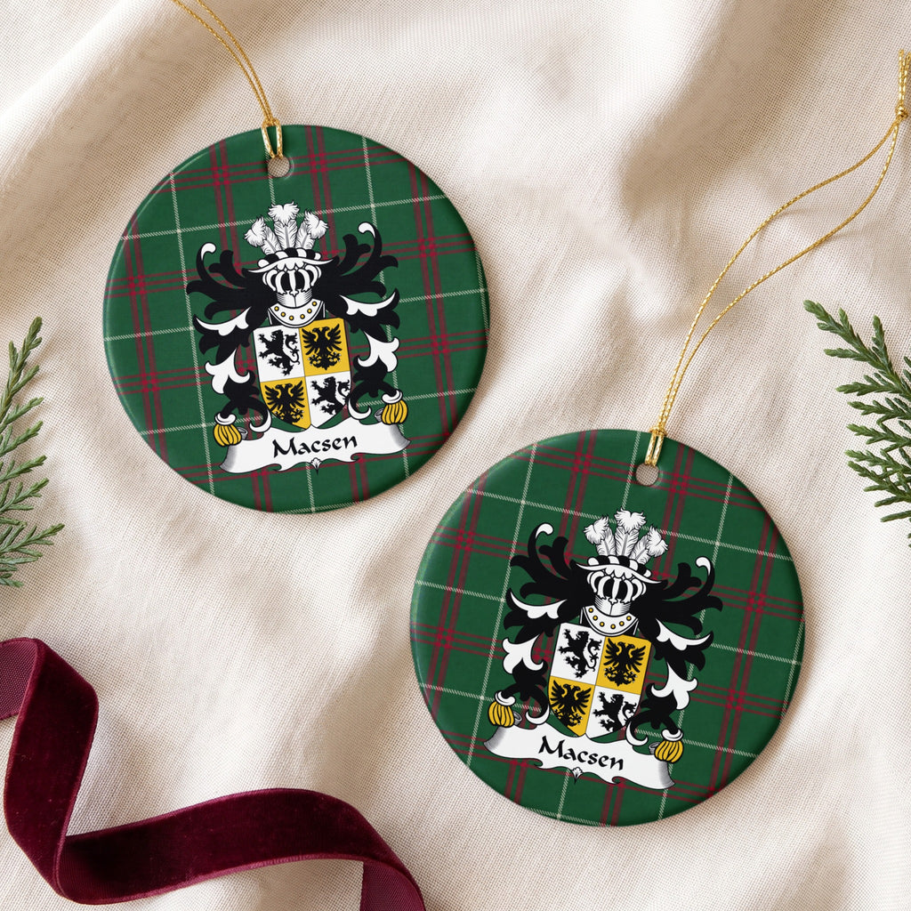 Two Macsen ornaments on cream fabric beside evergreen and burgundy ribbon, showing a quartered white-and-gold shield with black lions and eagles on Welsh National Tartan.