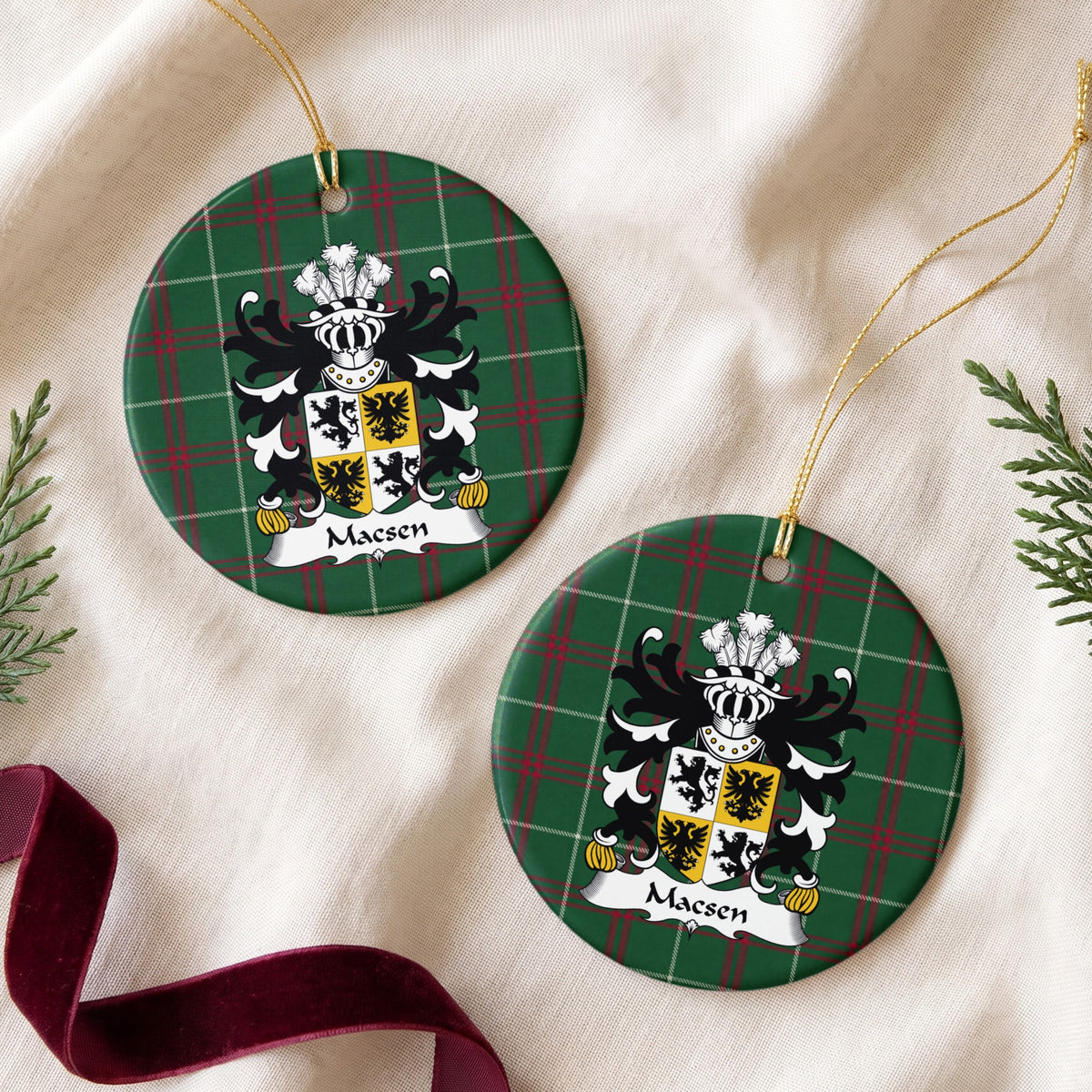 Two Macsen ornaments on cream fabric beside evergreen and burgundy ribbon, showing a quartered white-and-gold shield with black lions and eagles on Welsh National Tartan.