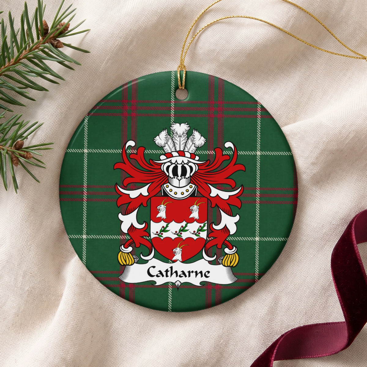 Catharne ornament on cream fabric with gold string, evergreen and burgundy ribbon; three white goat heads around a scalloped white band with leafy sprigs and red berries on a red shield on Welsh national tartan.