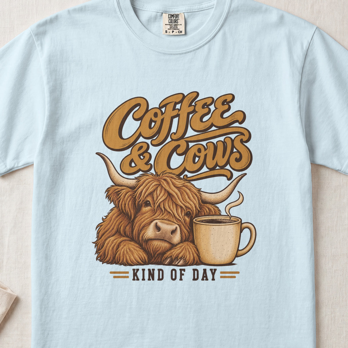 Coffee & Cows Kind of Day Highland cow design on a Chambray Comfort Colors 1717 T-shirt, flat lay