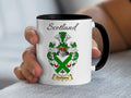 Hand-held white Clarkesone Scottish family shield mug with black handle, showing “Scotland” and a green-and-silver shield crossed by a green saltire with a red centre.