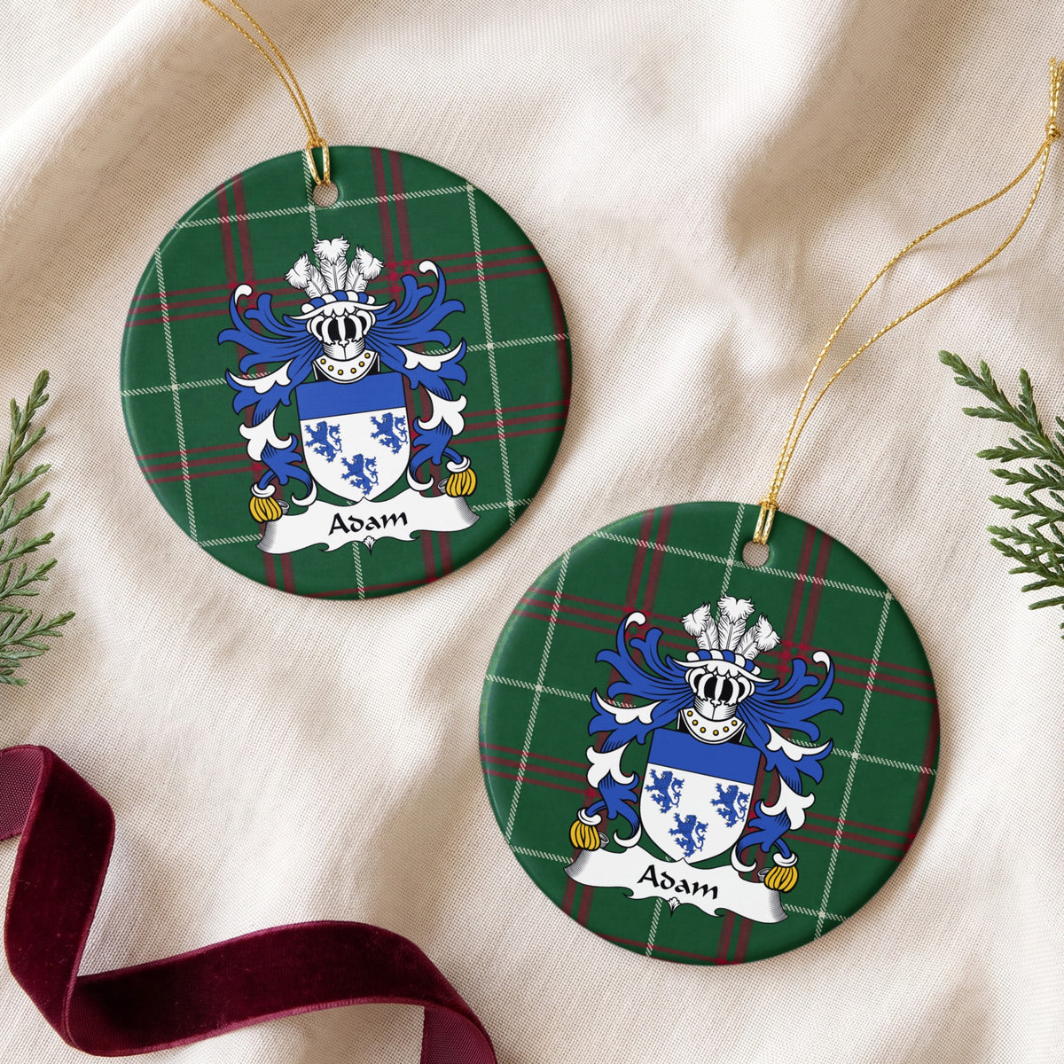 Two round ornaments showing the Adam family crest and surname on Welsh national tartan, arranged on cream fabric with gold cords, evergreen sprigs and burgundy ribbon.