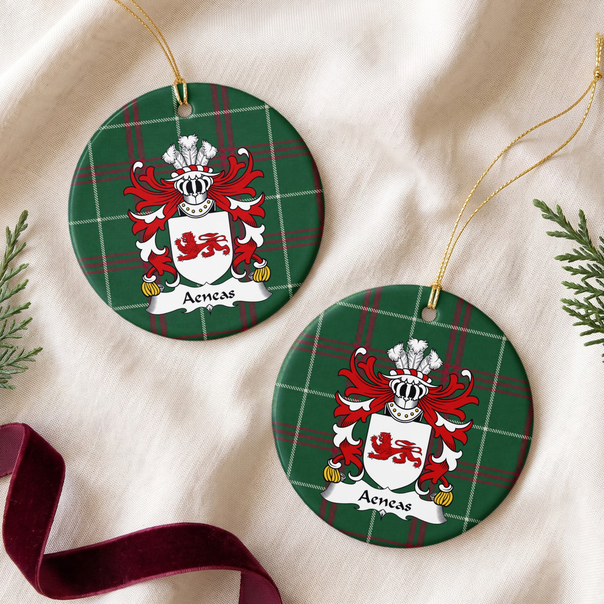 Two round ornaments showing the Aeneas family crest and surname on Welsh national tartan, arranged on cream fabric with gold cords, evergreen sprigs and burgundy ribbon.