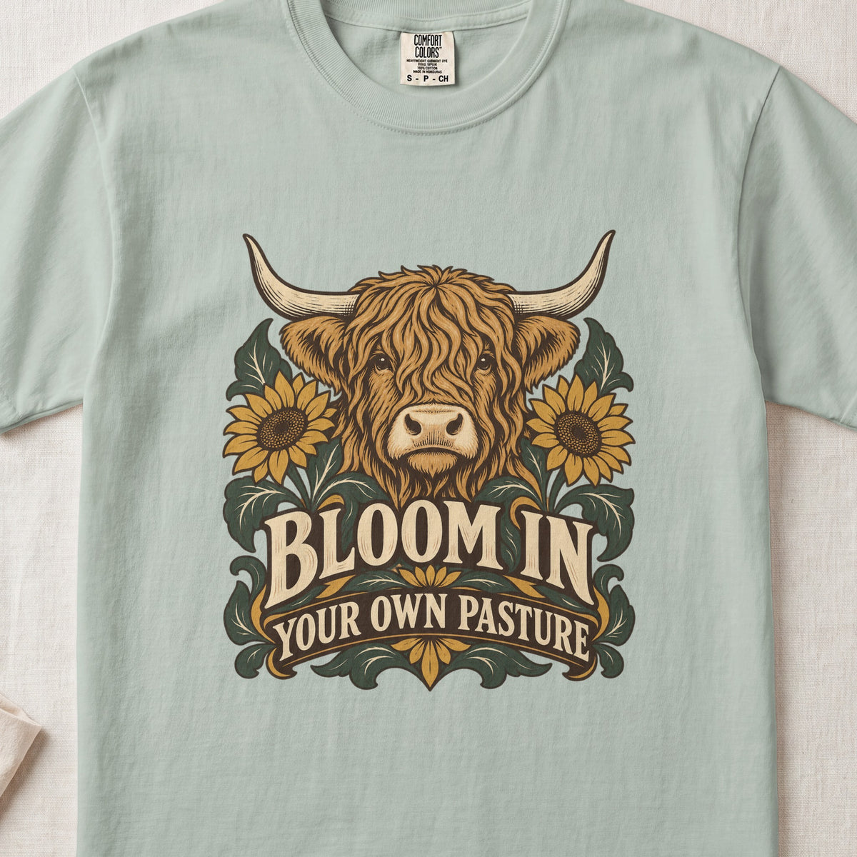 Bloom in Your Own Pasture Highland cow design on a Bay Comfort Colors 1717 T-shirt, flat lay