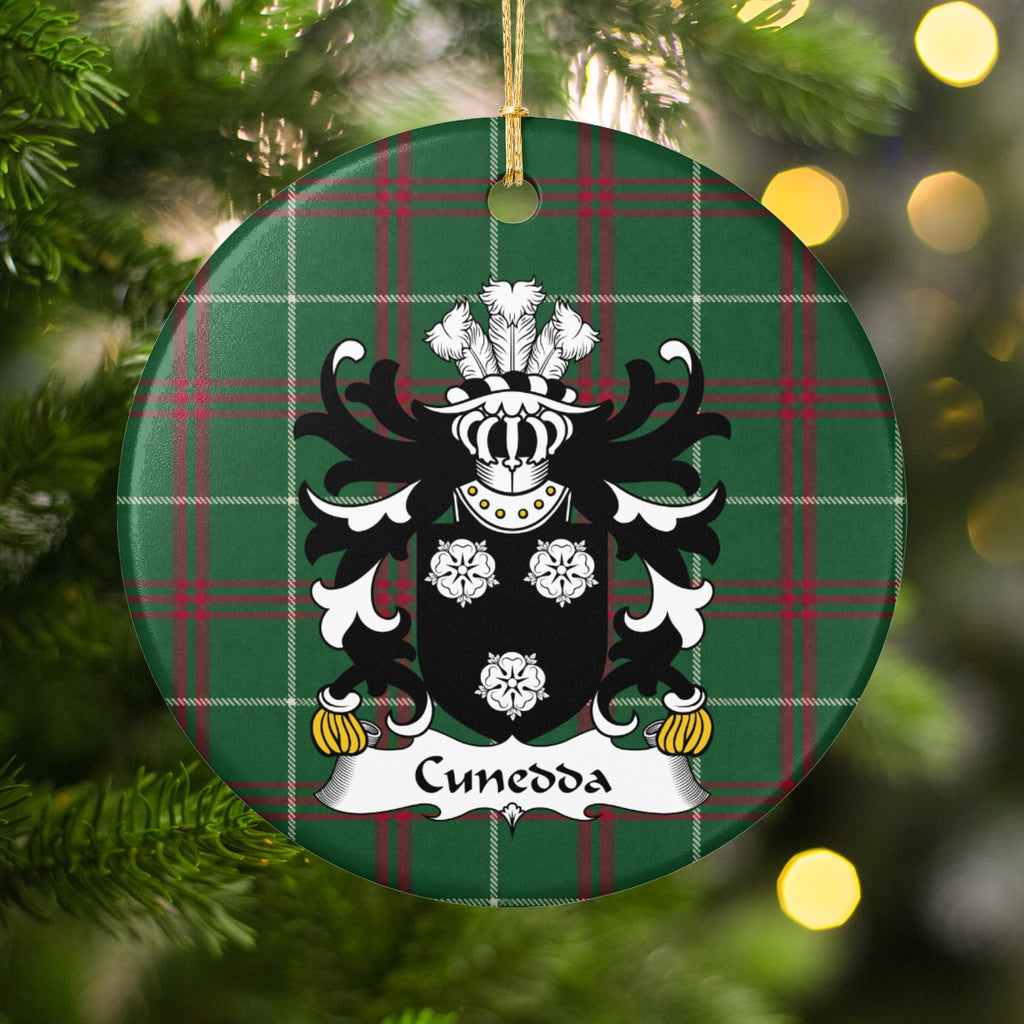 Cunedda ornament hanging among Christmas tree branches and warm lights; three white rose motifs on a black shield on Welsh national tartan.