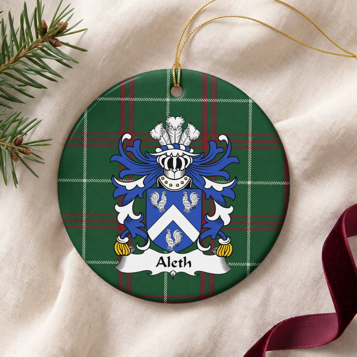 Aleth family crest and surname on a round Welsh national tartan ornament with a gold cord, resting on cream fabric beside an evergreen sprig and burgundy ribbon.