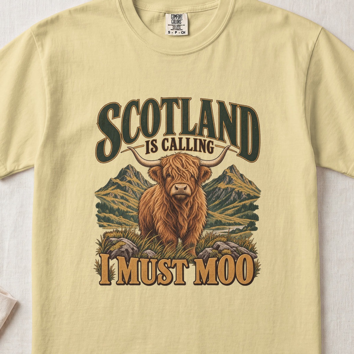 Scotland Is Calling, I Must Moo Highland cow design on a Butter Comfort Colors 1717 T-shirt, flat lay