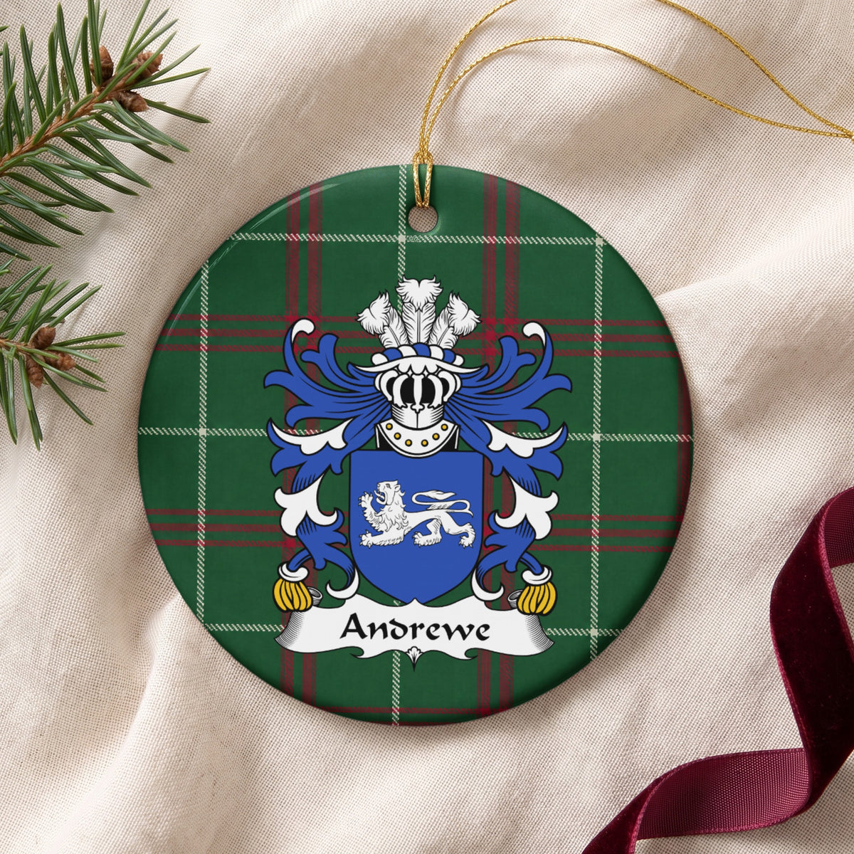 Andrewe family crest and surname on a round Welsh national tartan ornament with a gold cord, resting on cream fabric beside an evergreen sprig and burgundy ribbon.