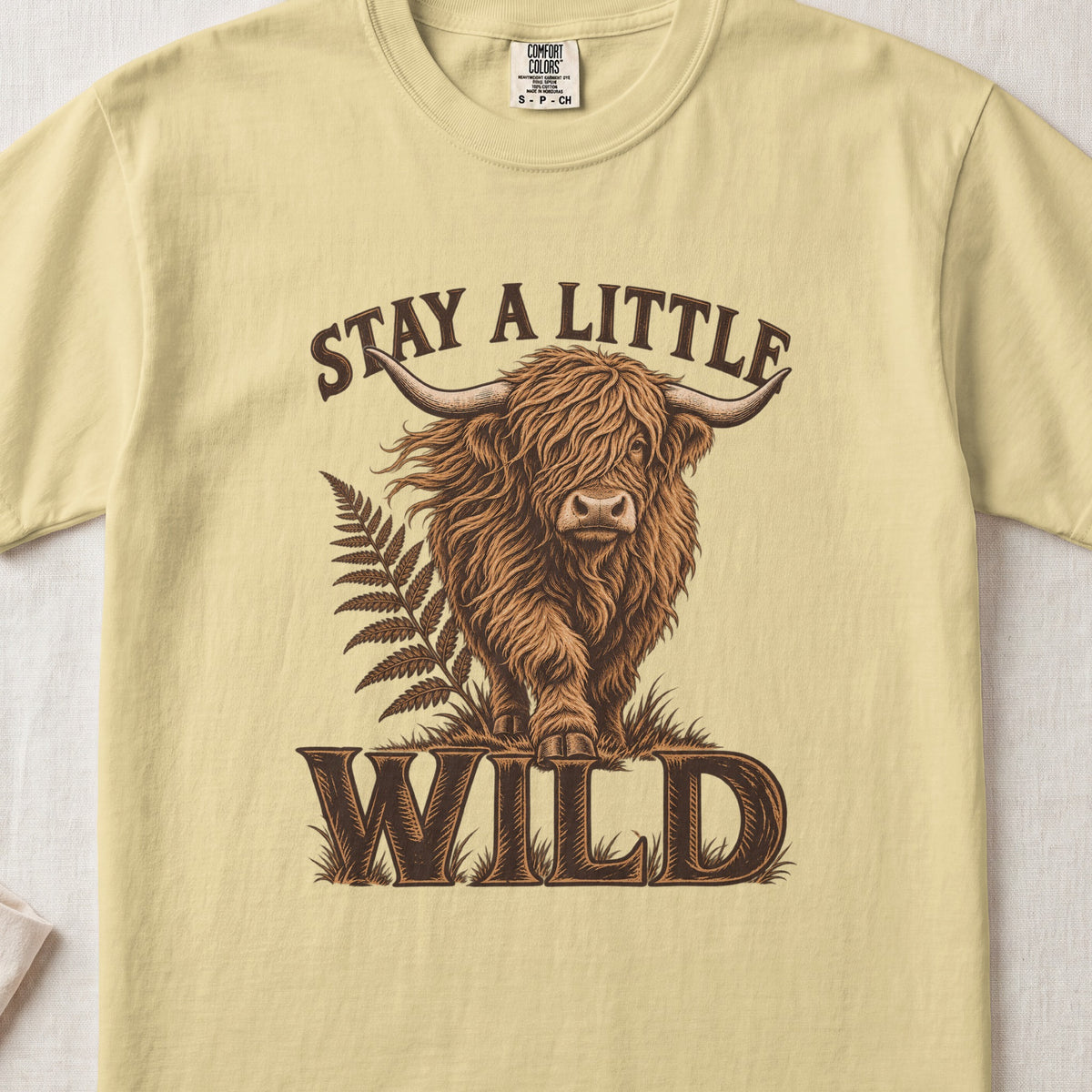 Stay a Little Wild Highland cow design on a Butter Comfort Colors 1717 T-shirt, flat lay