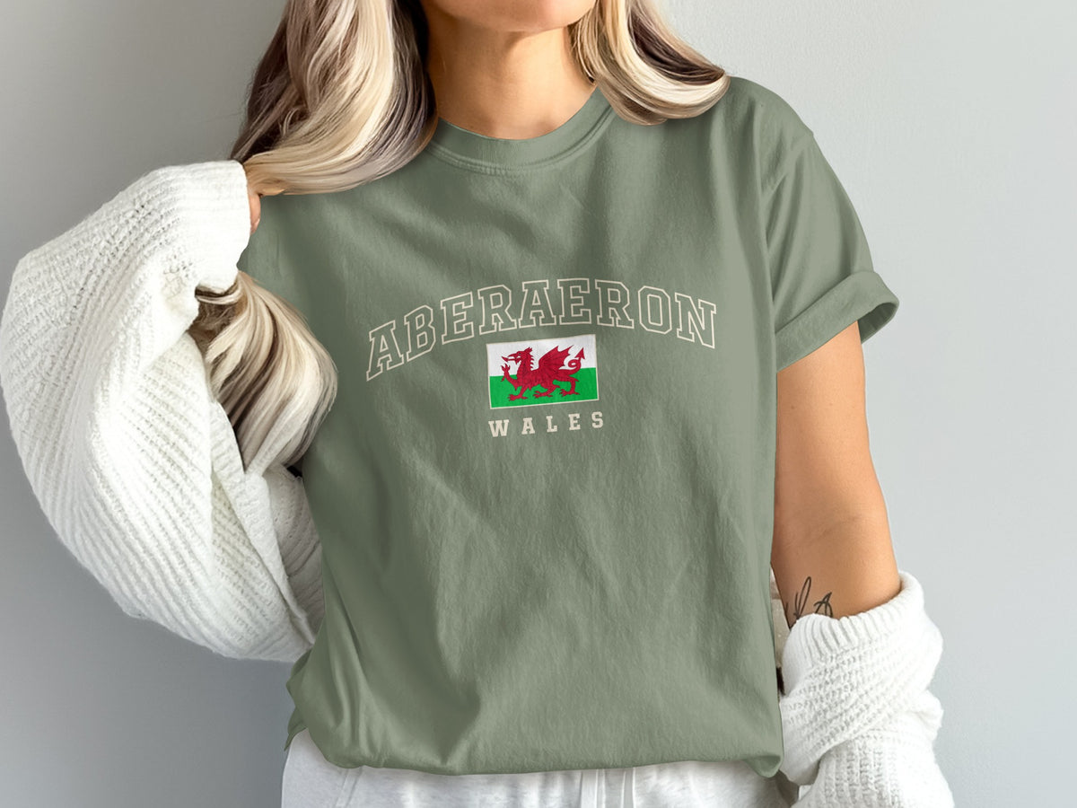 Model wearing a moss green Aberaeron Wales T-shirt with a Welsh dragon flag and a white cardigan draped off the shoulders.