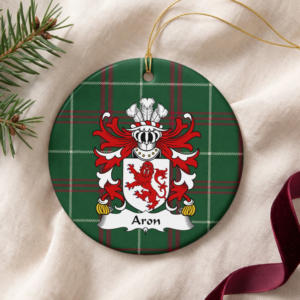 Aron family crest and surname on a round Welsh national tartan ornament with a gold cord, resting on cream fabric beside an evergreen sprig and burgundy ribbon.