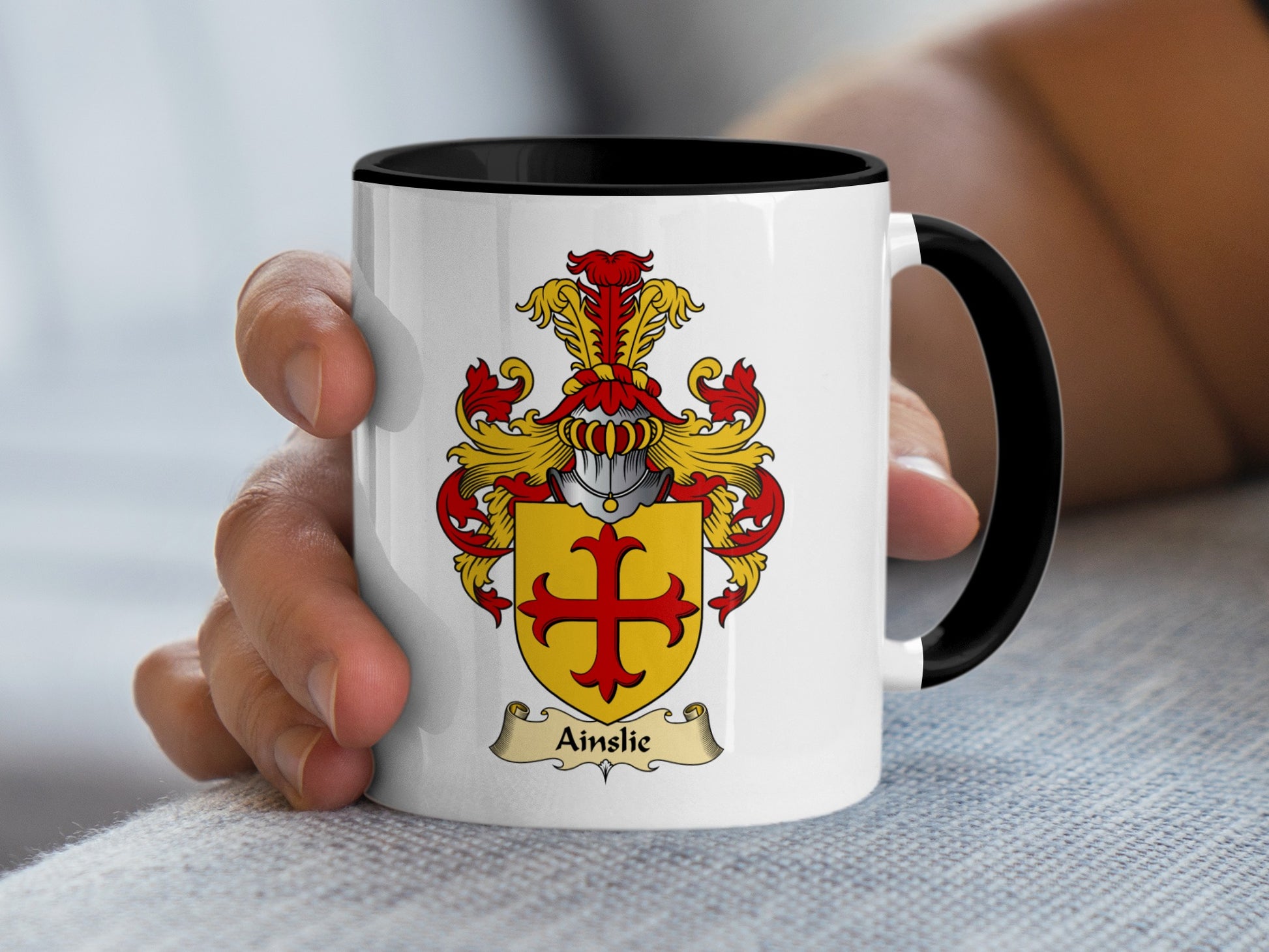 White mug with black interior held by a hand, featuring the Ainslie coat of arms.