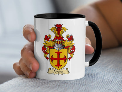 White mug with black interior held by a hand, featuring the Ainslie coat of arms.