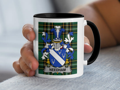 White mug with black rim featuring Needham family crest on plaid background.
