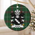 Arthal family crest and surname on a round Welsh national tartan ornament, hanging by a gold cord from a pale wooden branch with blurred Christmas lights behind it.