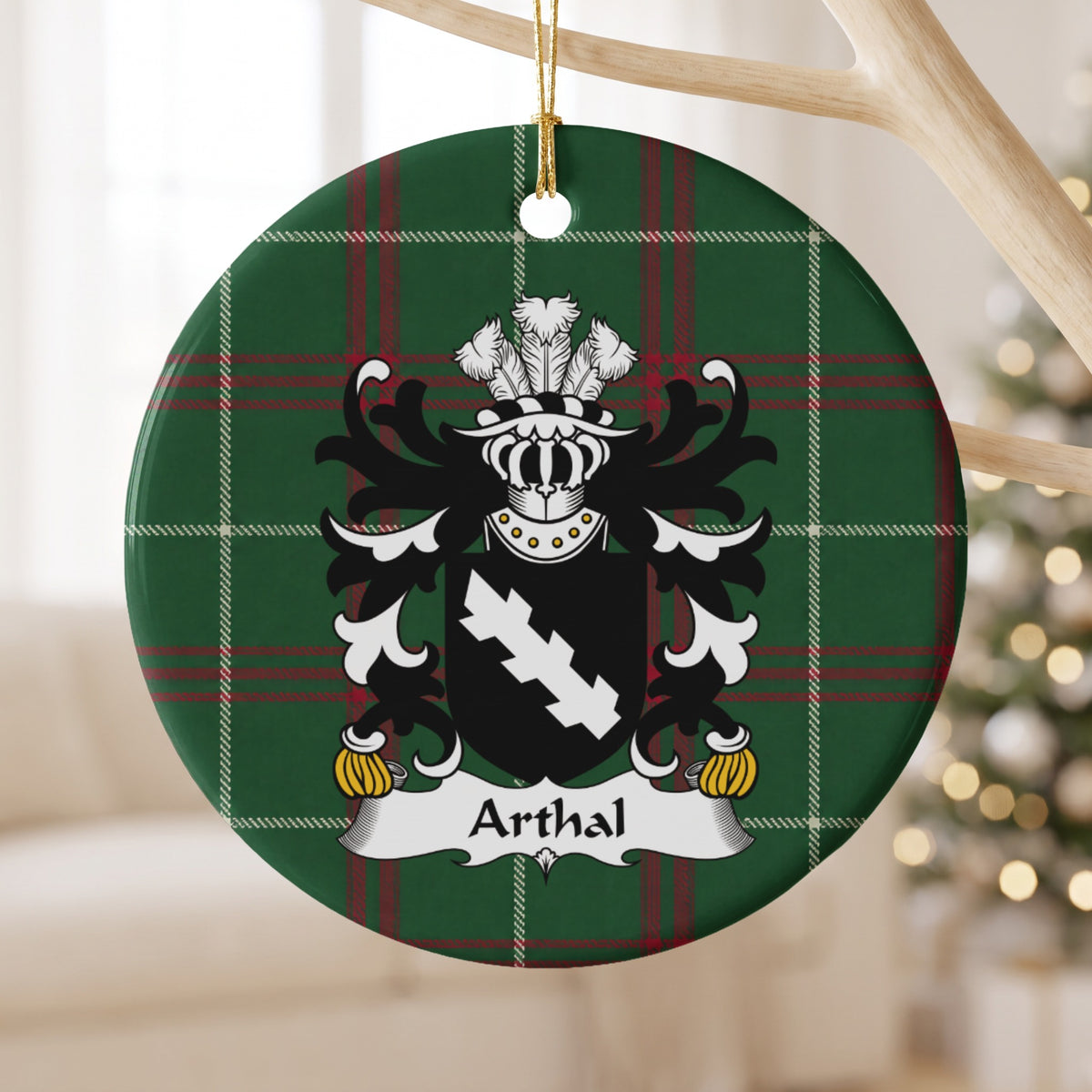 Arthal family crest and surname on a round Welsh national tartan ornament, hanging by a gold cord from a pale wooden branch with blurred Christmas lights behind it.