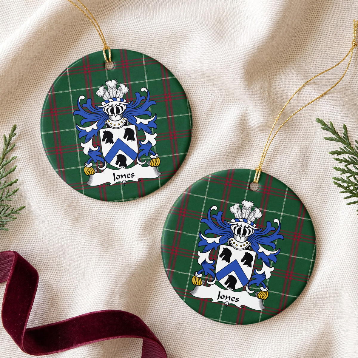 Two Jones Style 3 ornaments on cream fabric beside evergreen sprigs and burgundy ribbon, showing a white shield with a blue chevron and three black animal heads on Welsh national tartan.