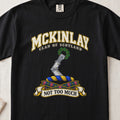 McKinlay Scottish clan T-shirt in Black, front view with an armored arm holding a green leafy wreath and motto banner.