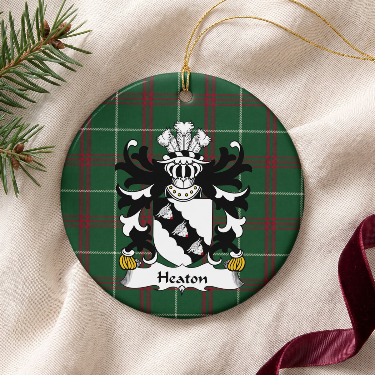 Heaton ornament on cream fabric beside evergreen and burgundy ribbon, with gold string; three white bull heads on a scalloped black diagonal band across a white shield.