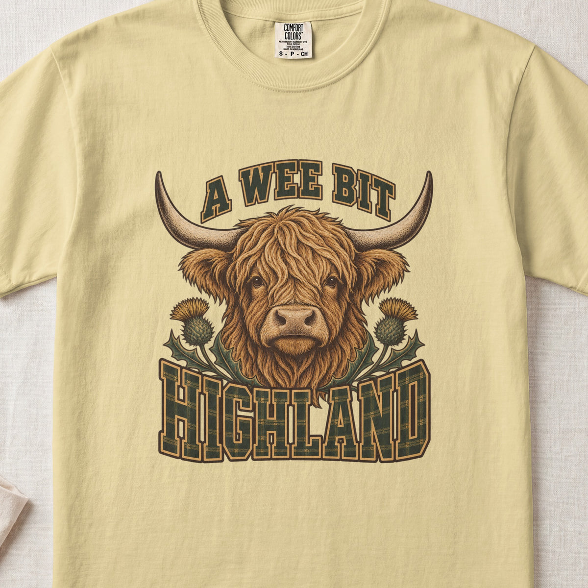 A Wee Bit Highland Highland cow design on a Butter Comfort Colors 1717 T-shirt, flat lay