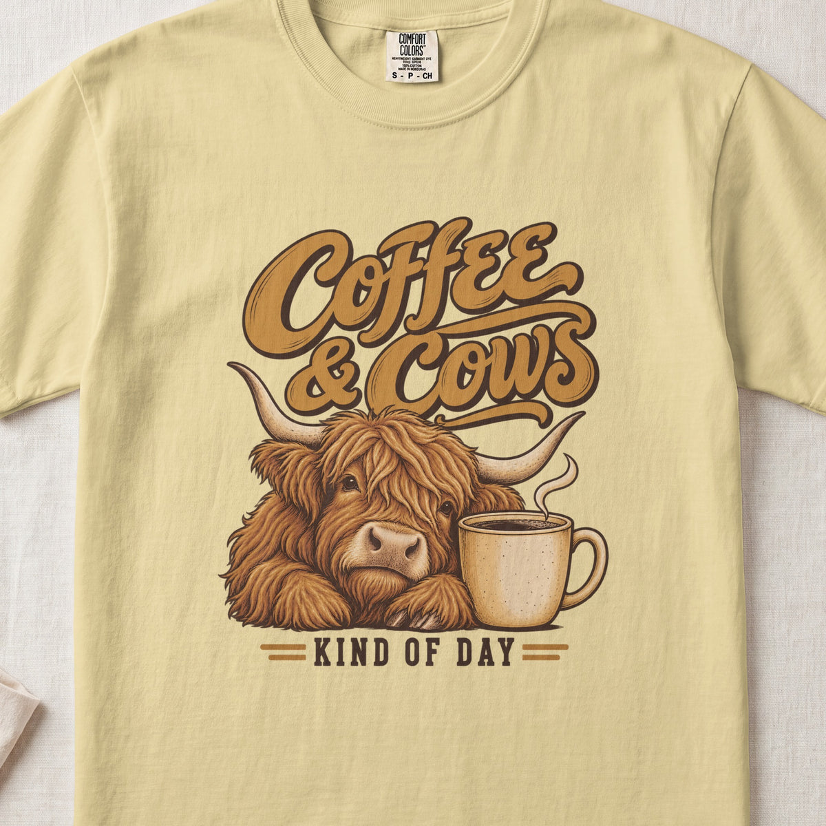 Coffee & Cows Kind of Day Highland cow design on a Butter Comfort Colors 1717 T-shirt, flat lay
