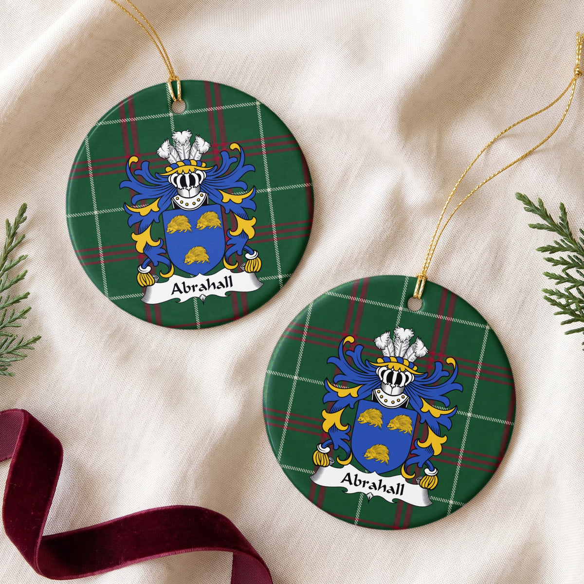 Two round ornaments showing the Abrahall family crest and surname on Welsh national tartan, arranged on cream fabric with gold cords, evergreen sprigs and burgundy ribbon.