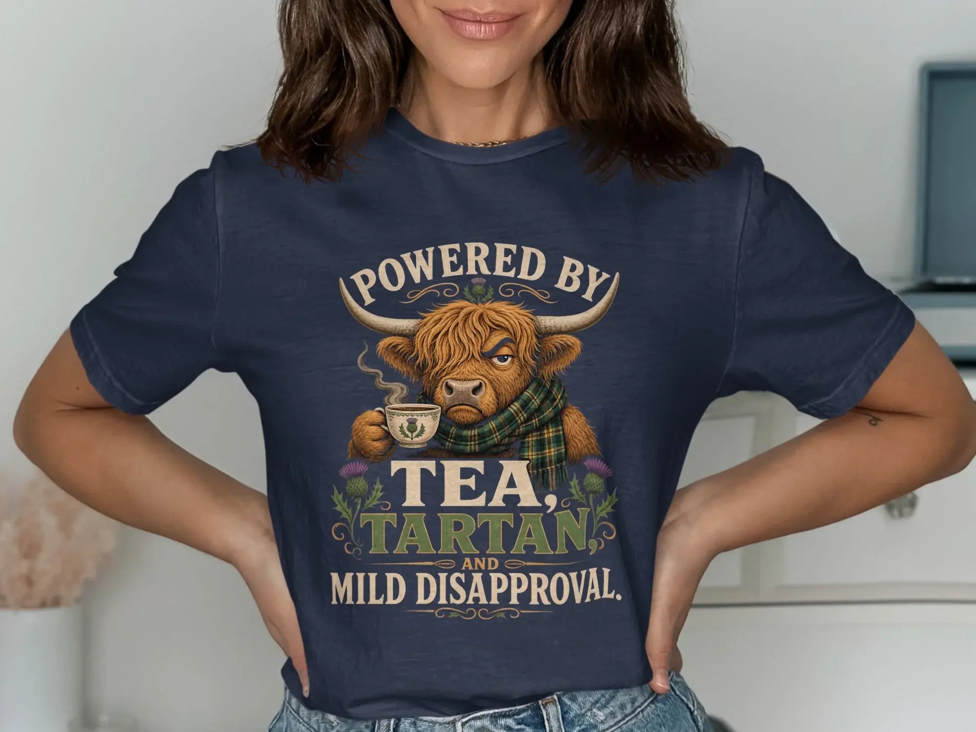 Navy t-shirt with Highland cow wearing scarf, holding tea, text reads: Powered by Tea, Tartan, and Mild Disapproval.