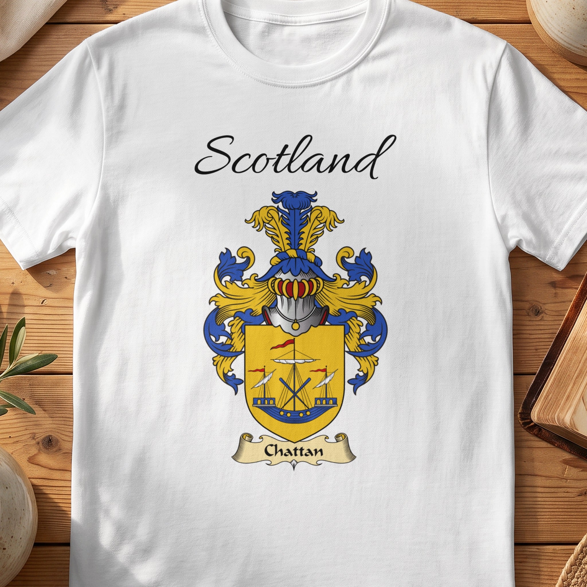 A white t-shirt featuring the Scotland Chattan clan crest with a ship design on a wooden table.