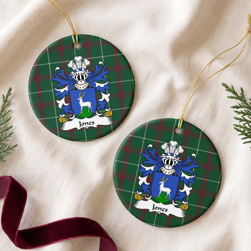 Two Jones Style 1 ornaments on cream fabric beside evergreen sprigs and burgundy ribbon, showing a blue shield with a white stag standing on a green mound on Welsh national tartan.