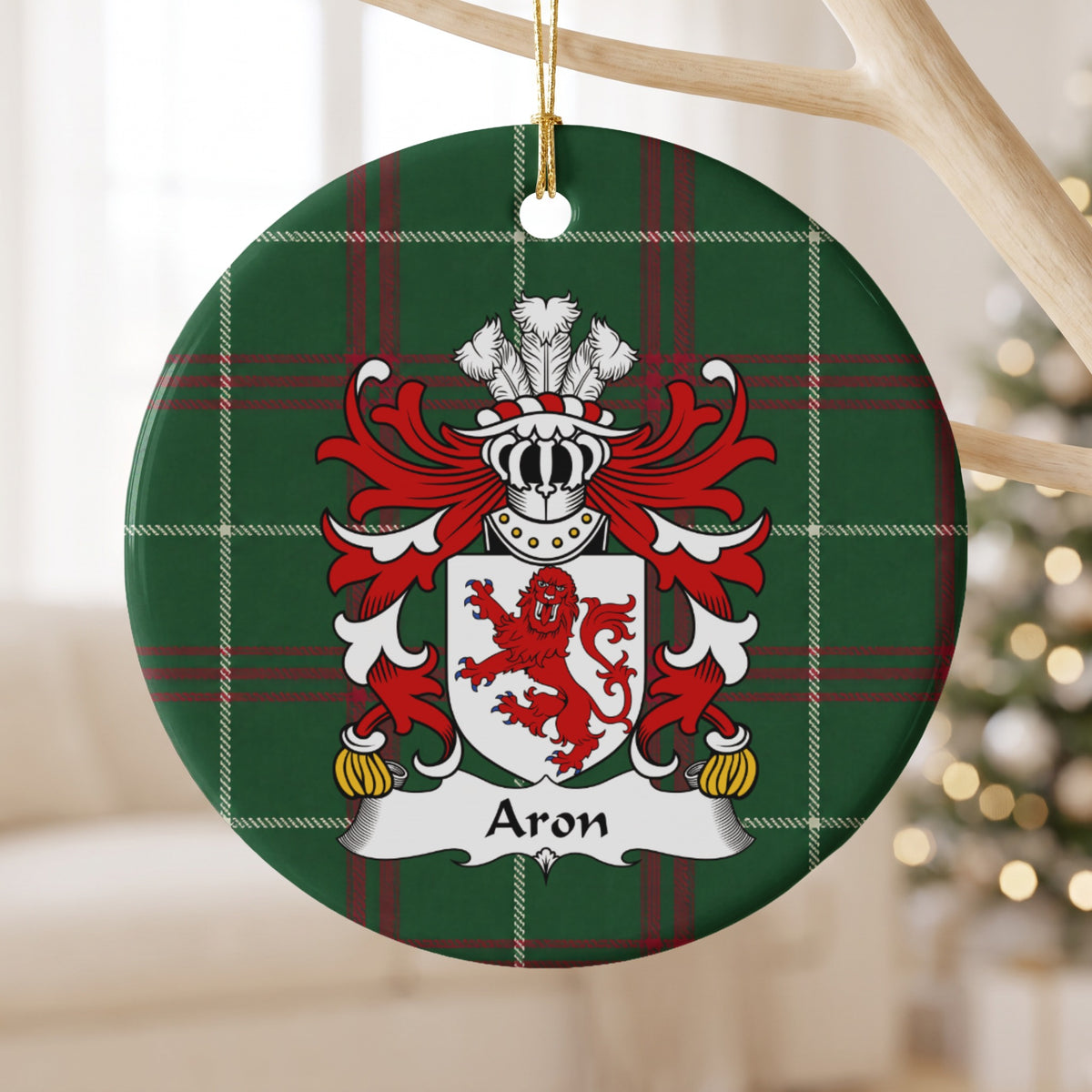 Aron family crest and surname on a round Welsh national tartan ornament, hanging by a gold cord from a pale wooden branch with blurred Christmas lights behind it.