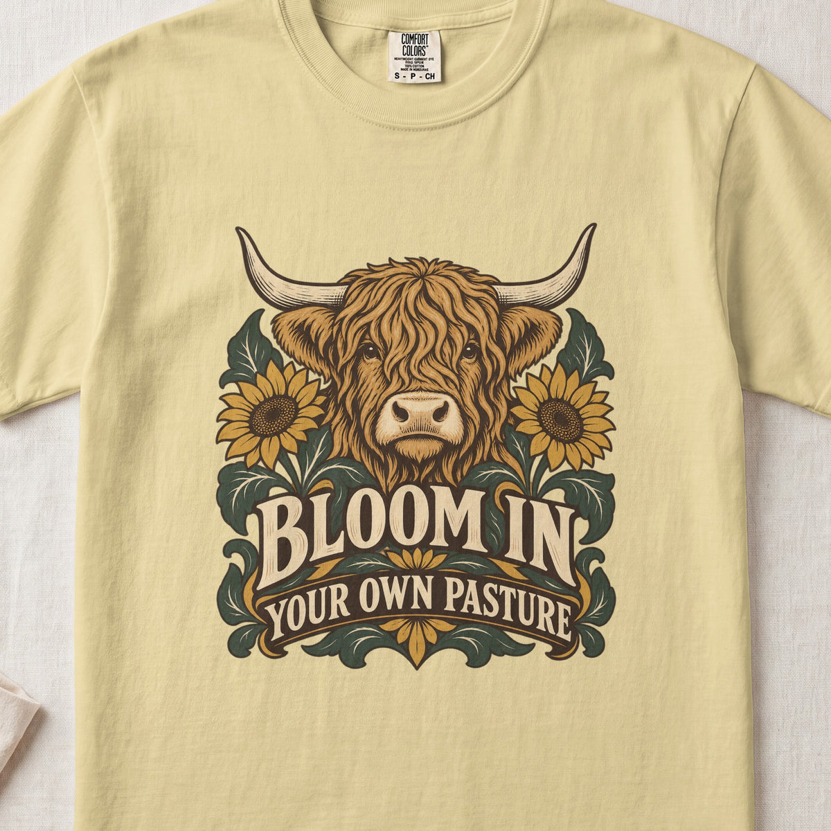 Bloom in Your Own Pasture Highland cow design on a Butter Comfort Colors 1717 T-shirt, flat lay