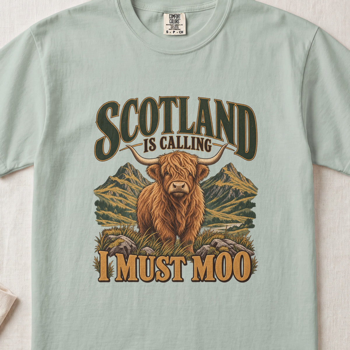 Scotland Is Calling, I Must Moo Highland cow design on a Bay Comfort Colors 1717 T-shirt, flat lay