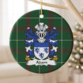 Adam family crest and surname on a round Welsh national tartan ornament, hanging by a gold cord from a pale wooden branch with blurred Christmas lights behind it.