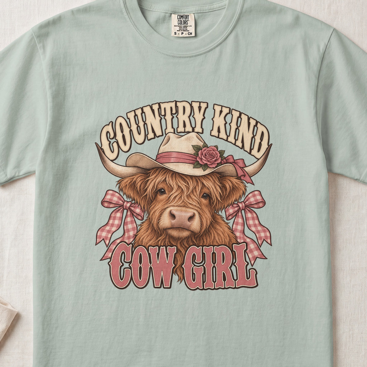 Country Kind Cow Girl Highland cow design on a Bay Comfort Colors 1717 T-shirt, flat lay
