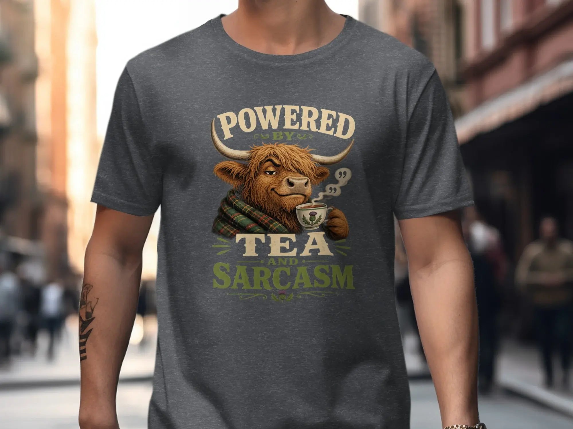 Dark grey t-shirt with a cartoon Highland cow wearing a tartan scarf, holding a mug, text "Powered by Tea and Sarcasm".
