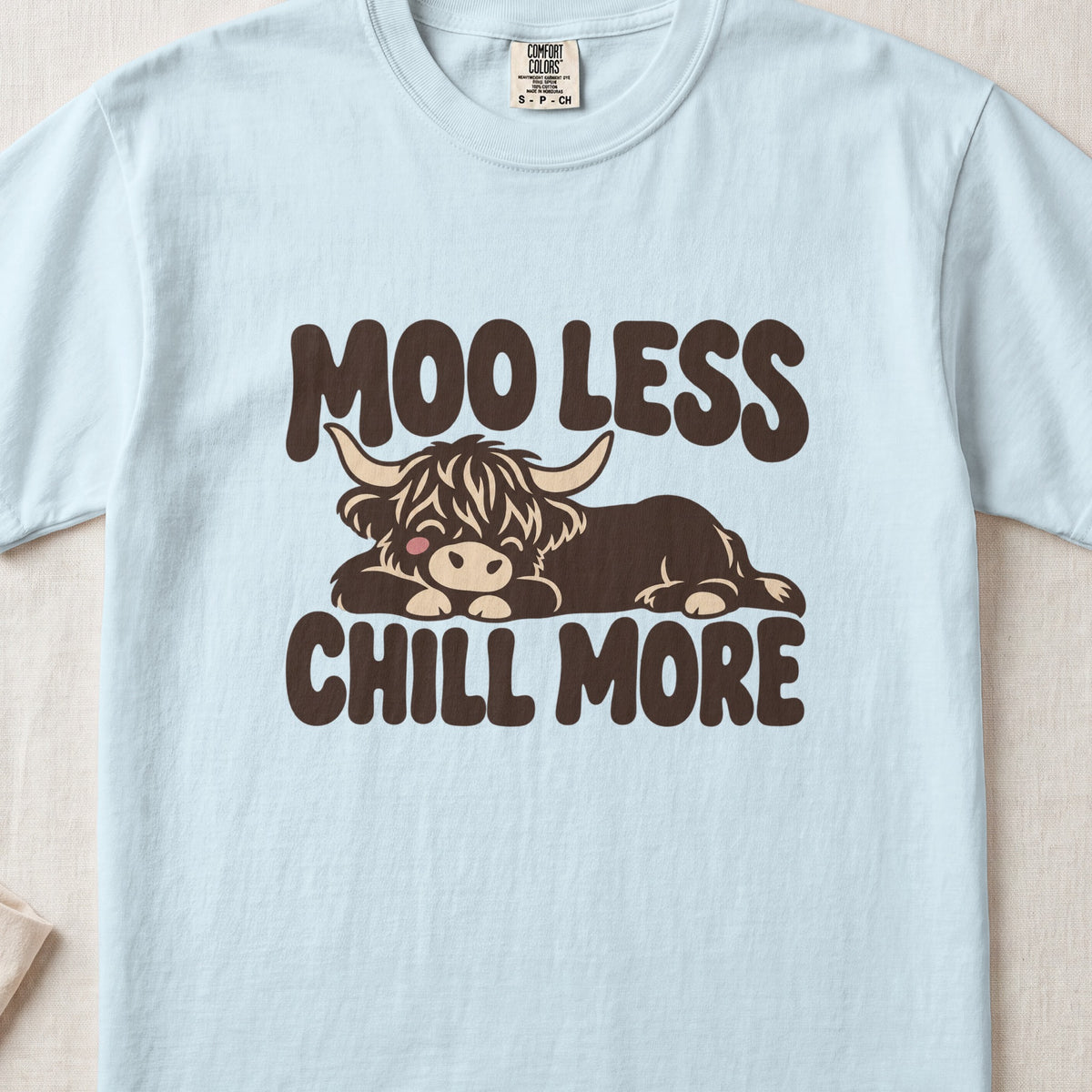 Moo Less, Chill More Highland cow design on a Chambray Comfort Colors 1717 T-shirt, flat lay