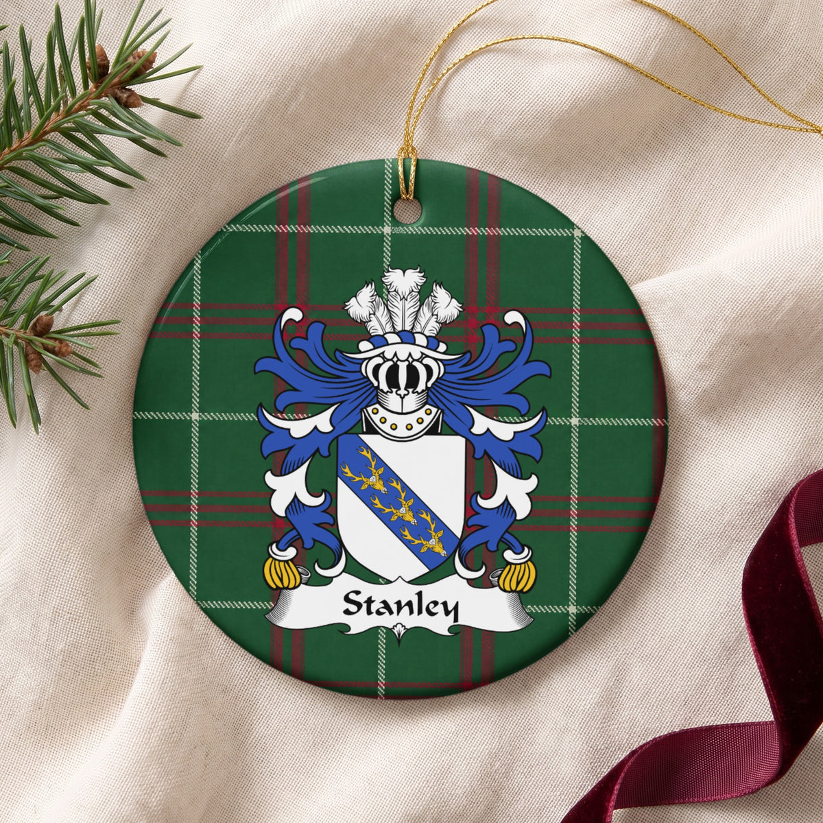 Stanley family crest ornament with Welsh national tartan background and gold cord, lying on cream fabric beside evergreen sprigs and burgundy ribbon.