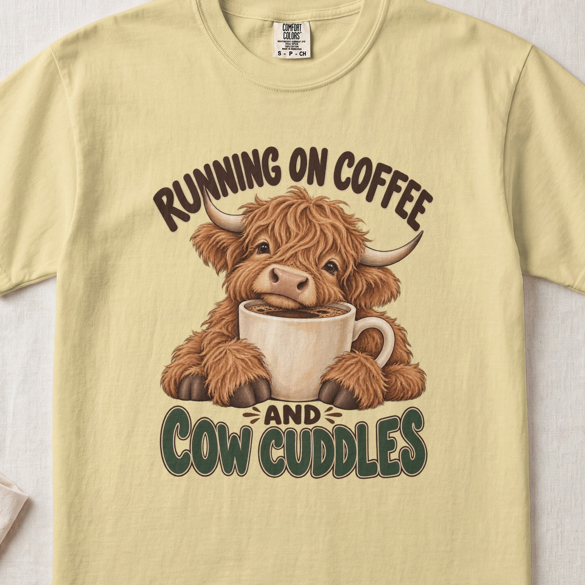 Running on Coffee & Cow Cuddles Highland cow design on a Butter Comfort Colors 1717 T-shirt, flat lay
