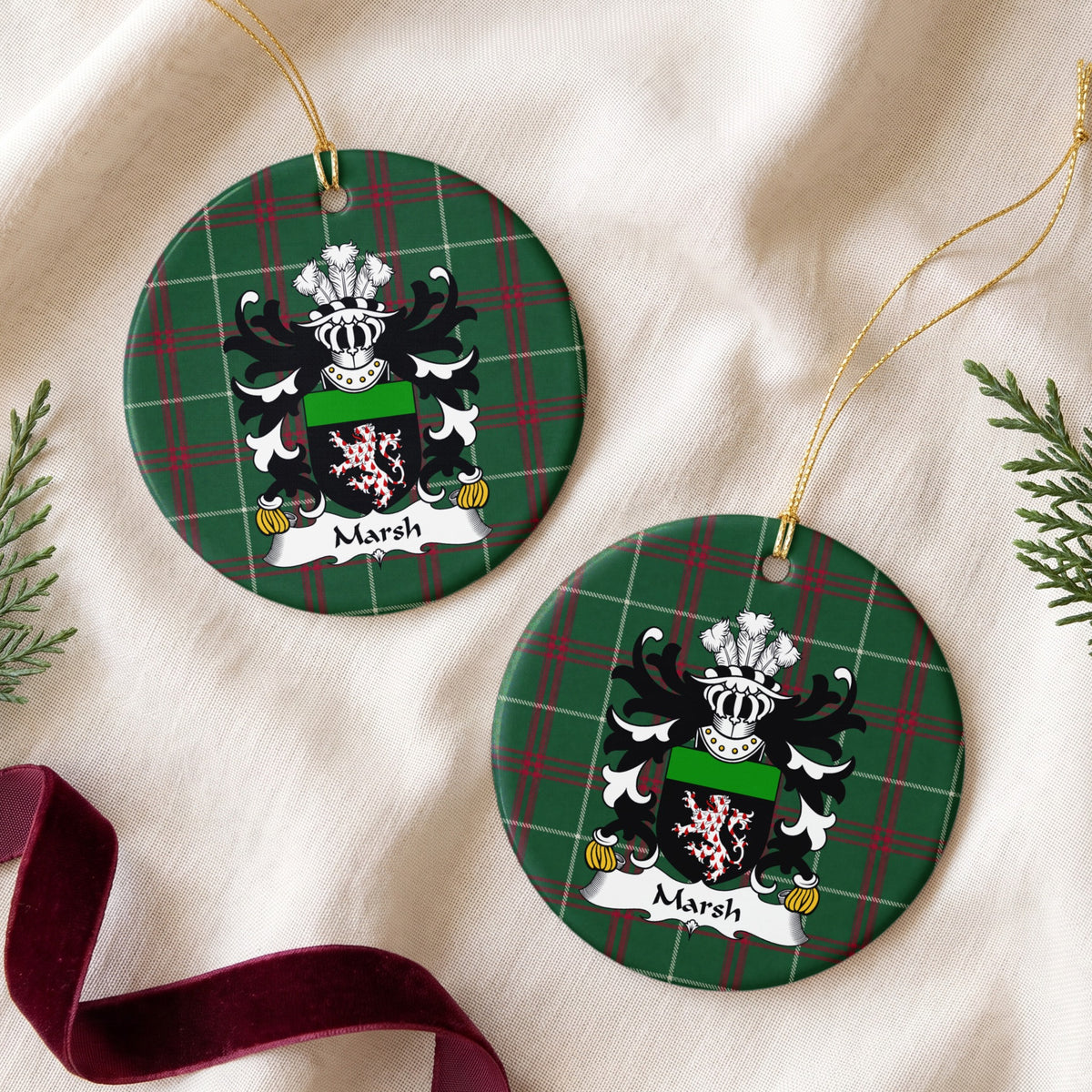 Two Marsh ornaments on cream fabric beside evergreen and burgundy ribbon, showing a red-and-white patterned lion on a black shield with a green top band on Welsh National Tartan.