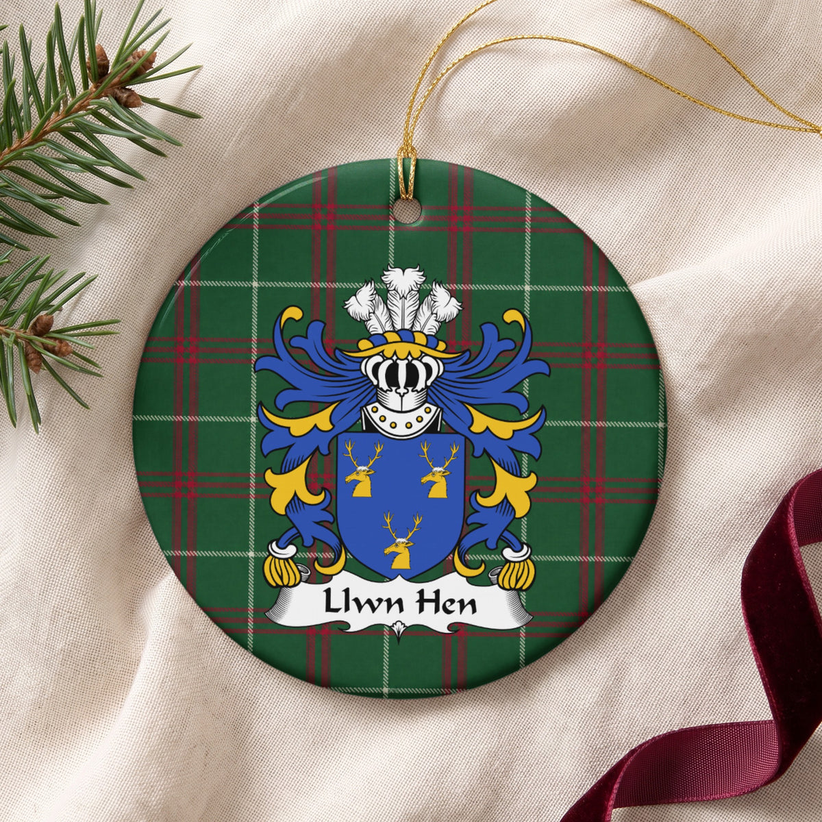 Llwn Hen ornament on cream fabric with gold hanging string, evergreen and burgundy ribbon; a blue shield with three gold stag heads on Welsh national tartan.