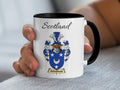 Hand-held white Arbuthnott Scottish family shield mug with black handle, showing “Scotland” and a blue-and-silver shield with a large crescent and small stars.