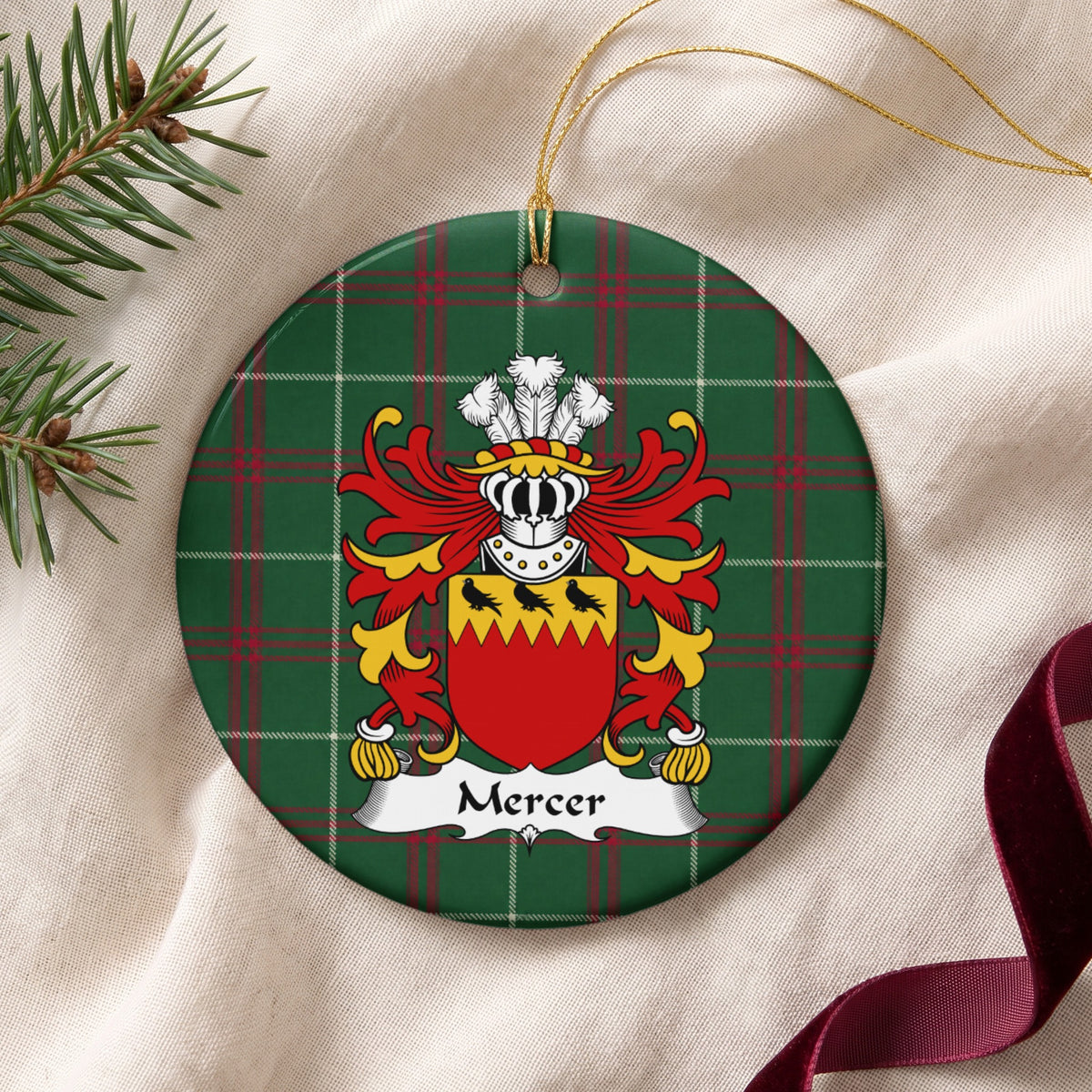 Mercer ornament on cream fabric with gold string, evergreen and burgundy ribbon; three black birds on a jagged gold band above a red shield on Welsh national tartan.