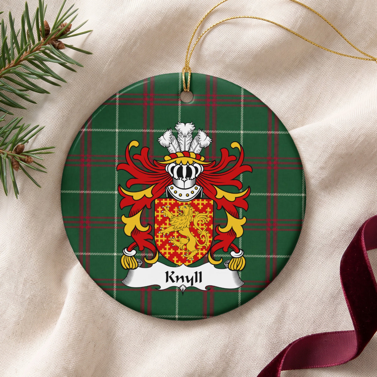 Knyll ornament on cream fabric with gold hanging string, evergreen and burgundy ribbon; a red shield with a gold lion surrounded by small gold crosses on Welsh national tartan.