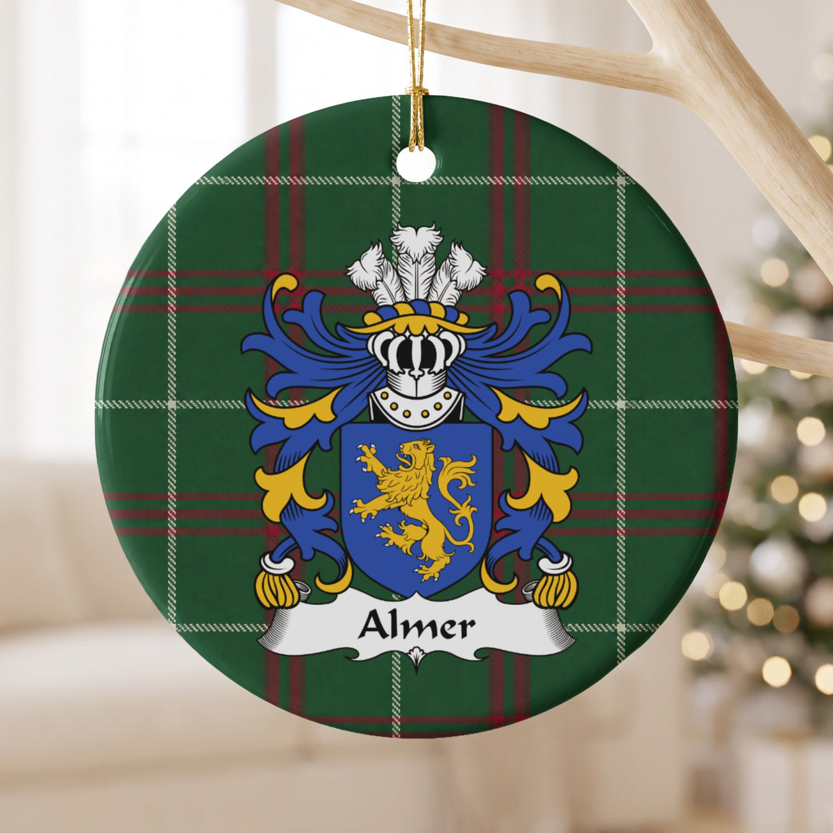 Almer family crest and surname on a round Welsh national tartan ornament, hanging by a gold cord from a pale wooden branch with blurred Christmas lights behind it.
