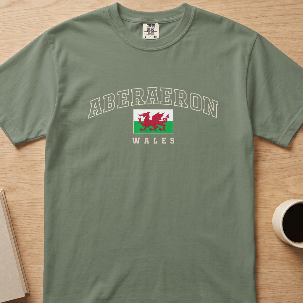 Moss green Aberaeron Wales T-shirt laid flat, with cream collegiate outline lettering and a red Welsh dragon flag.